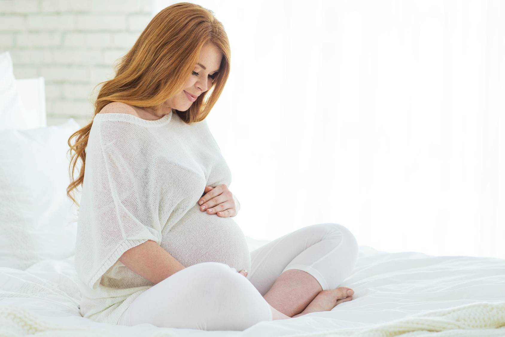 Ciąża po 40-tce - jak się dobrze przygotować do późnego macierzyństwa i jak pomóc sobie zajść w późną ciążę? Rudowłosa kobieta w zaawansowanej ciąży w białym luźnym ubraniu siedzi na łóżku na białej pościeli.