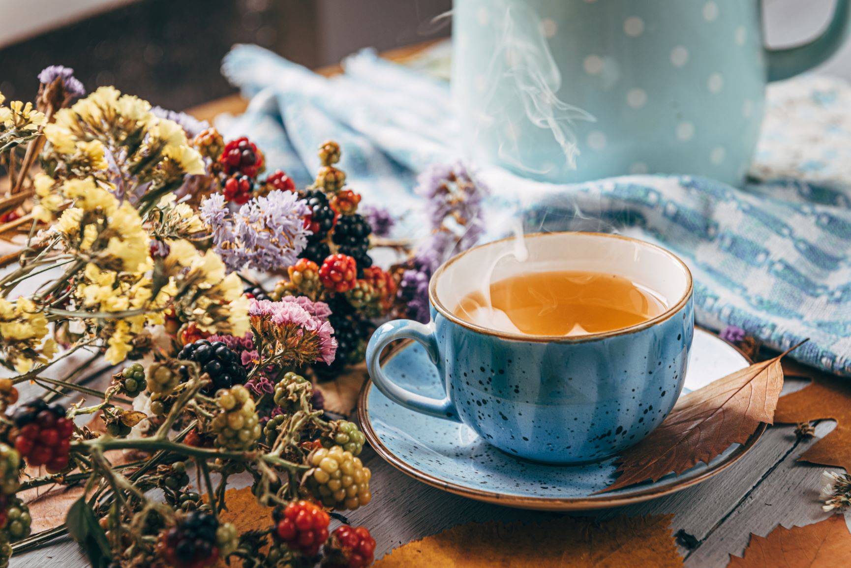 Herbata na kaszel - jak ją przyrządzić? Przepis na herbatę ziołową na kaszel. Niebieska filiżanka gorącej herbaty stoi na drewnianym stole obok bukietu ususzonych kwiatów.