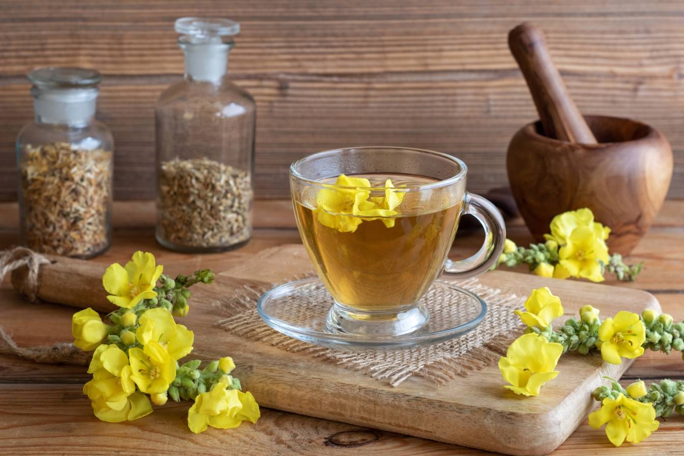 Jakie właściwości lecznicze ma dziewanna? Napar z dziewanny w szklanej filiżance na drewnianym stole, wokół leżą kwiaty dziewanny.