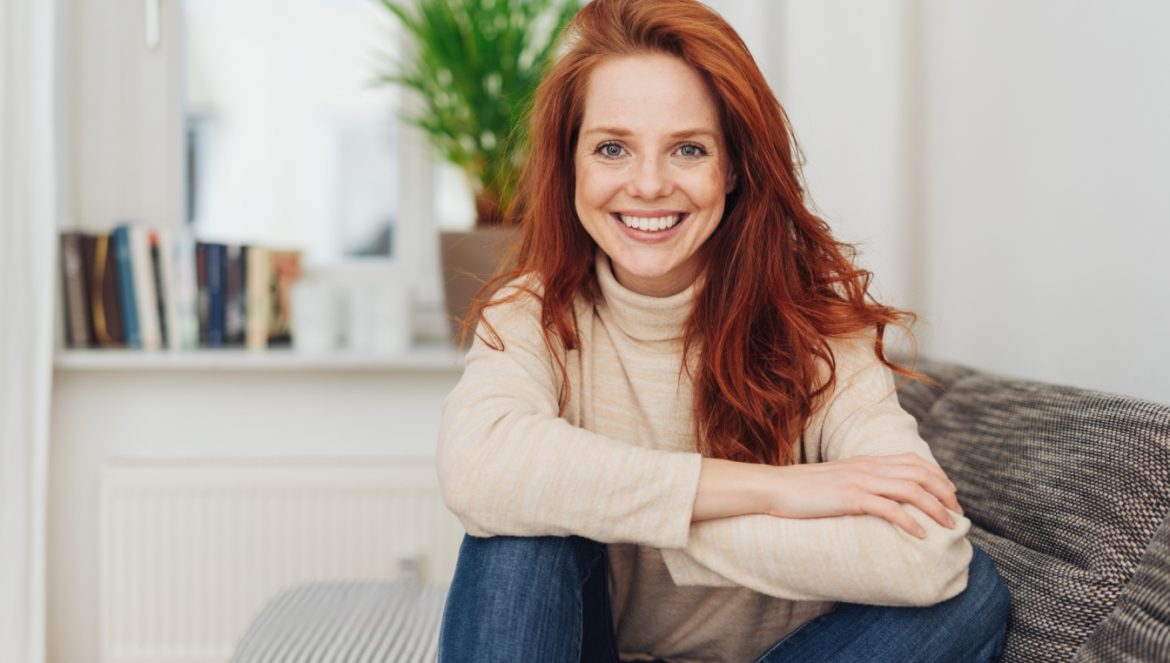 Homeopatia na czas rekonwalescencji - jakie leki homeopatyczne powinien zażywać rekonwalescent? Młoda rudowłosa dziewczyna siedzie na kanapie w salonie w ciepłym golfie i uśmiecha się do aparatu.