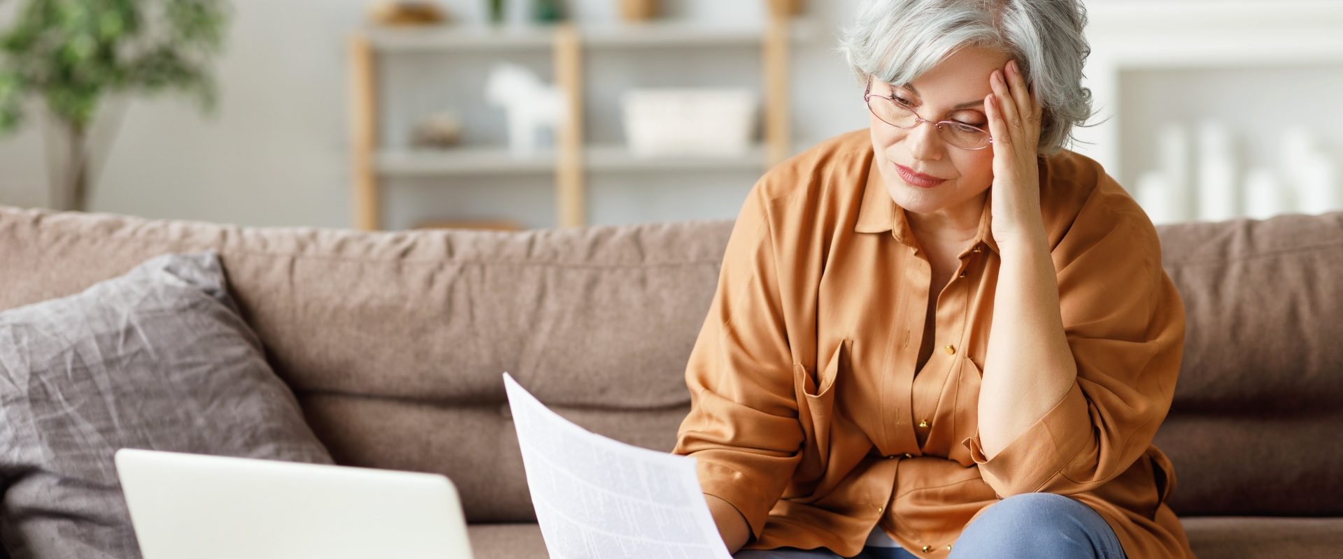 Leki homeopatyczne dla zestresowanych. Starsza zestresowana kobieta czyta ważne dokumenty, siedząc przed laptopem na kanapie w domu.