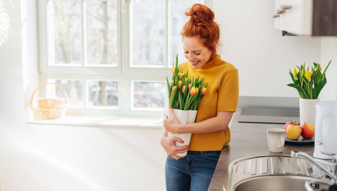 Homeopatia na syndrom wiosennego zmęczenia. Jakie leki homeopatyczne warto brać na przedwiośniu, czy czujemy, że dopadło nas wiosenne przesilenie? Młoda rudowłosa kobieta w żółtej bluzce stoi w kuchni obejmując bukiet świeżych tulipanów.