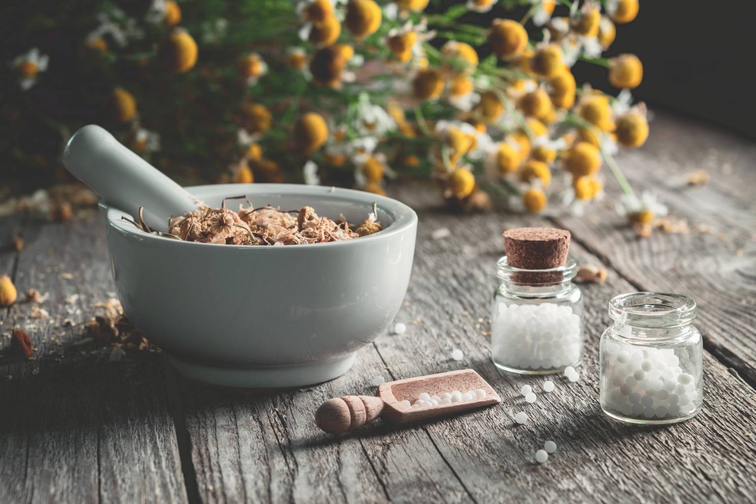 Leki homeopatyczne na przeziębienie. W białym moździerzu leżą suszone zioła, w tle bukiet rumianku, obok stoją fiolki z lekami homeopatycznymi.