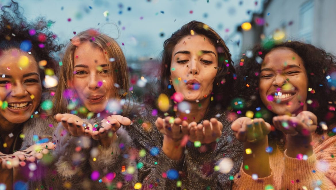 Hormony szczęścia: dopamina, serotonina, endorfina. Jak je podkręcić? Cztery uśmiechnięte dziewczyny zdmuchują z dłoni confetti.