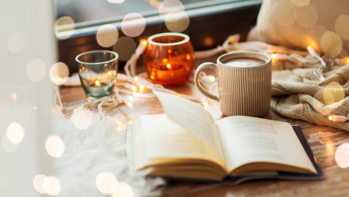 Jaki jest przepis na szczęście? Czym jest hygge, lagom, fika, sisu i cosagach? Zdjęcie przytulnego kącika - na parapecie leży kubek z herbatą, otwarta książka, zapalone świece i lampki dekoracyjne.