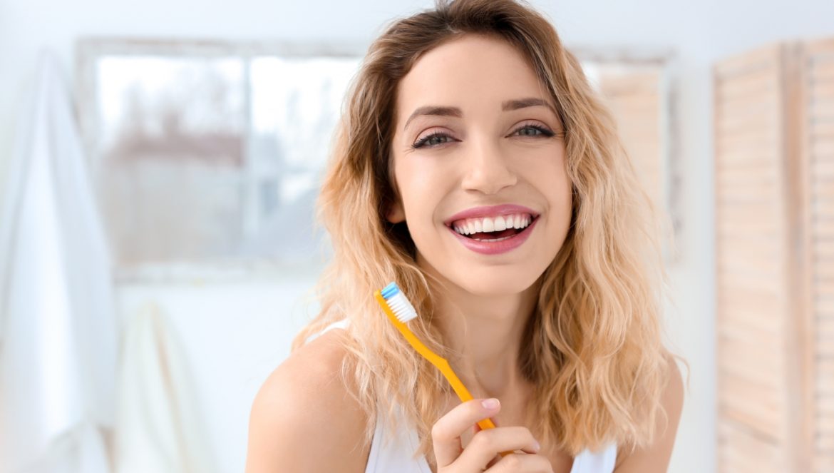 Szczoteczka do zębów - jaką wybrać? Czym się różni szczoteczka soniczna od elektrycznej i manualnej? Jak prawidłowo myć zęby? Młoda uśmiechnięta kobieta z pięknymi białymi zębami tryma w ręku manualną szczoteczkę do zębów.