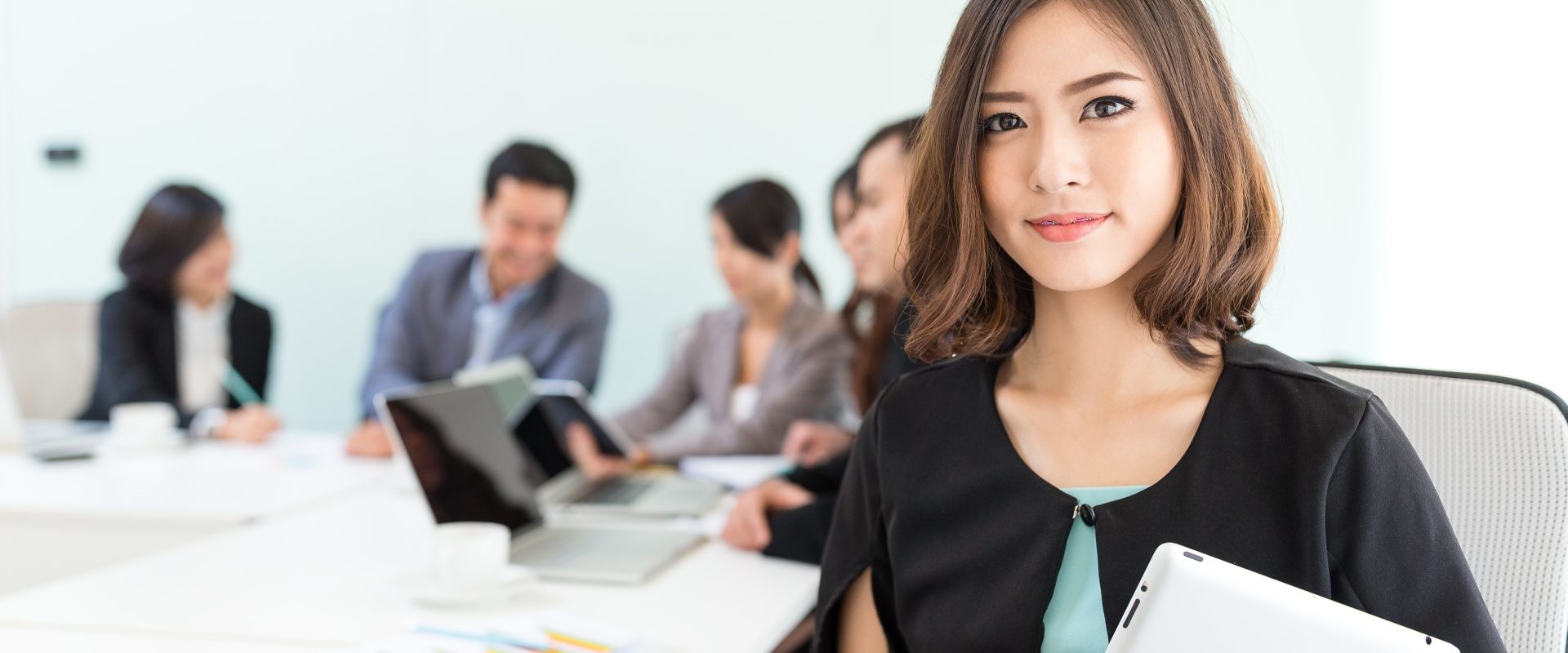 Nunchi - jak odnieść sukces po koreańsku? Piękna Azjatka patrzy w kamerę, a za nią siedzą współpracownicy na spotkaniu biznesowym.