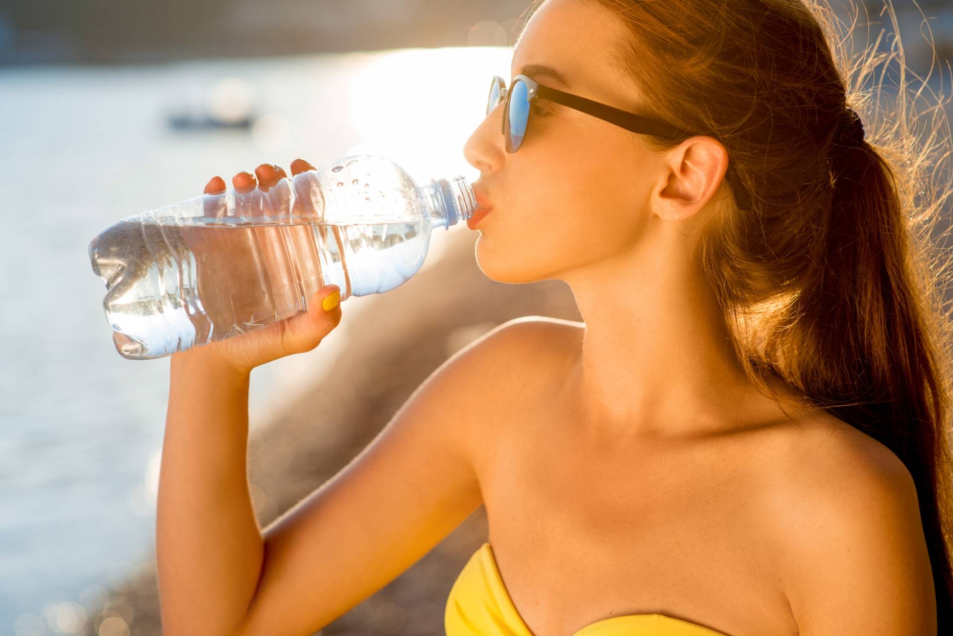 Ile wody dziennie pić? Co pić w upały? Młoda opalona kobieta w okularach przeciwsłonecznych, żółtym kostiumie kąpielowym, z włosami związanymi w kucyk, pije wodę z plastikowej butelki.