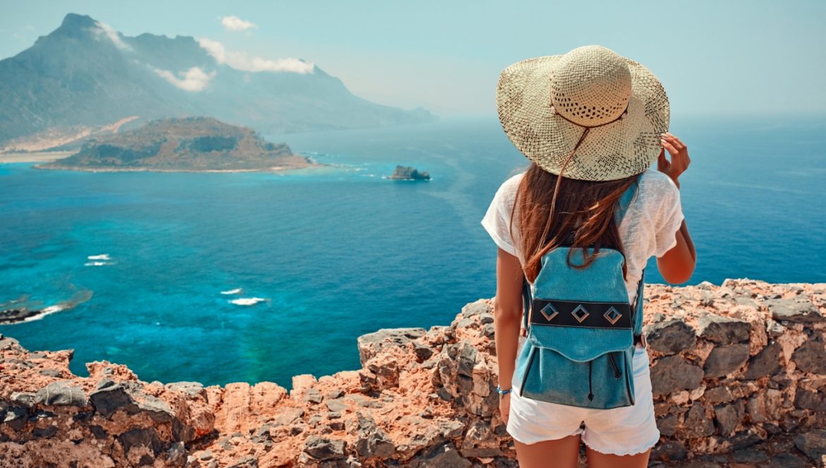 6 zasad jak być eko w podróży - jak zmnieszyć swój ślad węglowy na wakacjach? Dziewczyna w słomkowym kapeluszu i plecaku stoi na skarpie i podziwia panoramę - w tle ocean i skały.