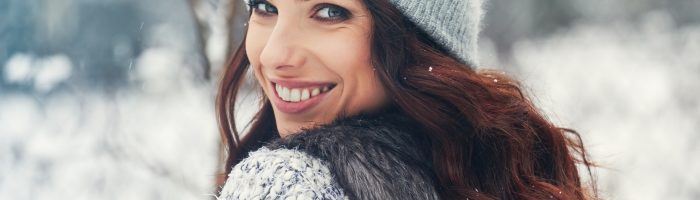 Naturalne sposoby na zapalenie zatok. Kobieta z długimi brązowymi włosami w szarej czapce, swetrze i bezrękawniku obraca się przez ramię, uśmiechając się do aparatu.