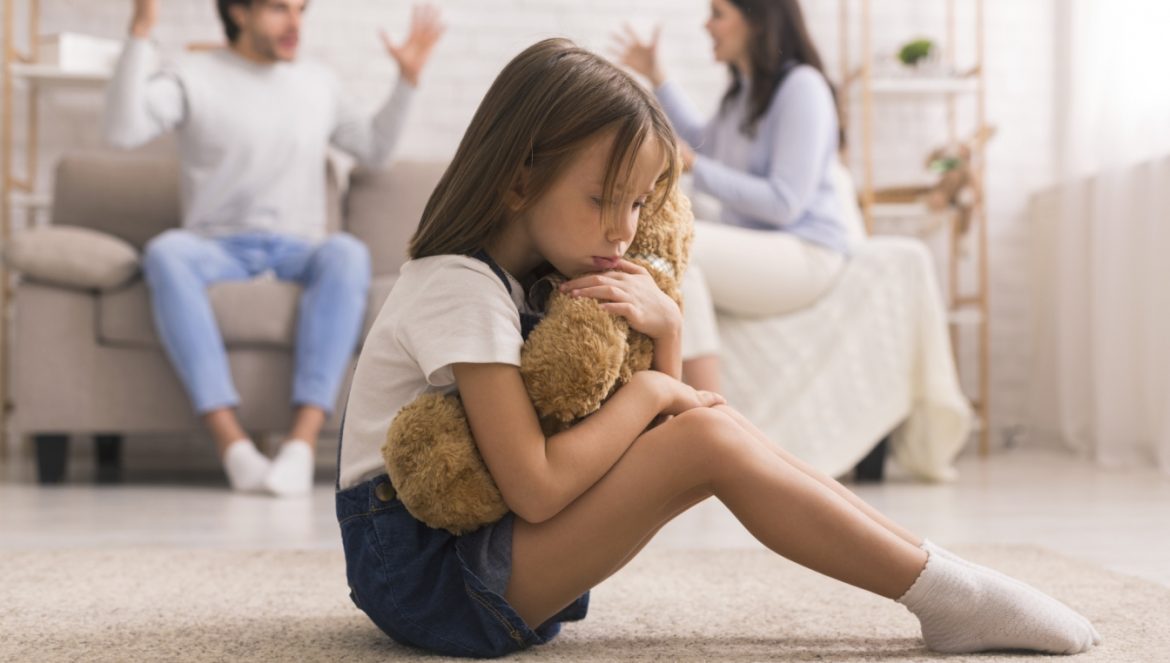 Jak rozpoznać depresję u dziecka? Objawy depresji u dzieci. Smutna dziewczynka przytula misia siedząc na podłodze w salonie w domu, podczas gdy rodzice kłócą się na kanapie.