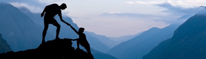 Jak pomóc osobie z depresją, w rozpaczy i kryzysie psychicznym? Osoba stojąca na skale wyciąga rękę i pomaga wspiąć się współtowarzyszowi wspinaczki - granatowa kolorystyka.