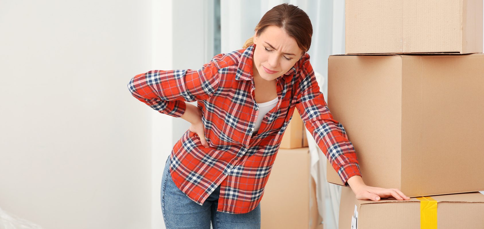 Jak prawidłowo podnosić ciężar? Kobieta trzyma się za kręgosłup lędźwiowy podczas przenoszenia kartonów.