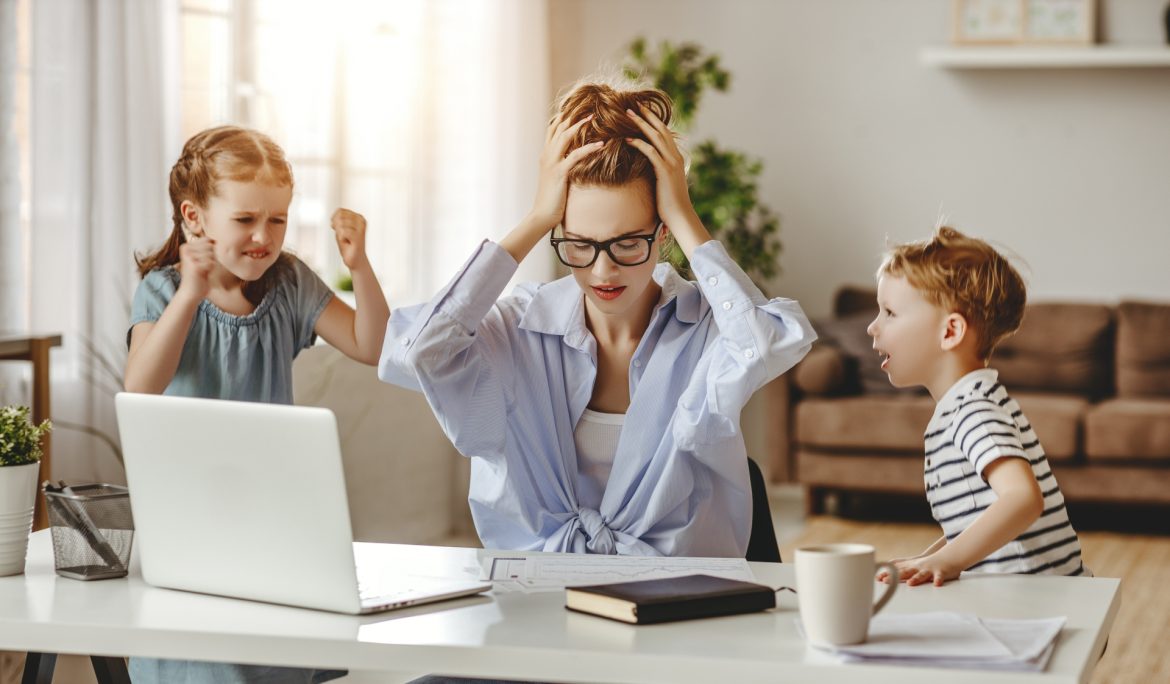 Jak radzić sobie ze stresem w XXI wieku? Zestresowana kobieta łapie się za głowę, siedząc przy laptopie, podczas gdy jej dzieci nie dają jej pracować.