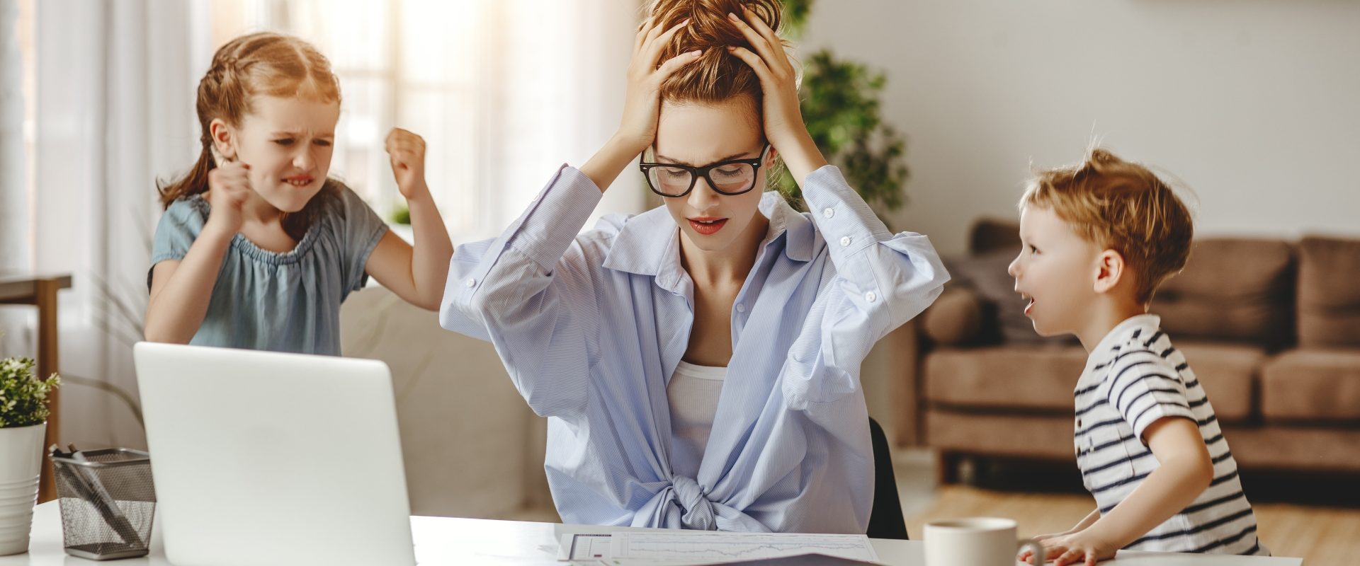 Jak radzić sobie ze stresem w XXI wieku? Zestresowana kobieta łapie się za głowę, siedząc przy laptopie, podczas gdy jej dzieci nie dają jej pracować.