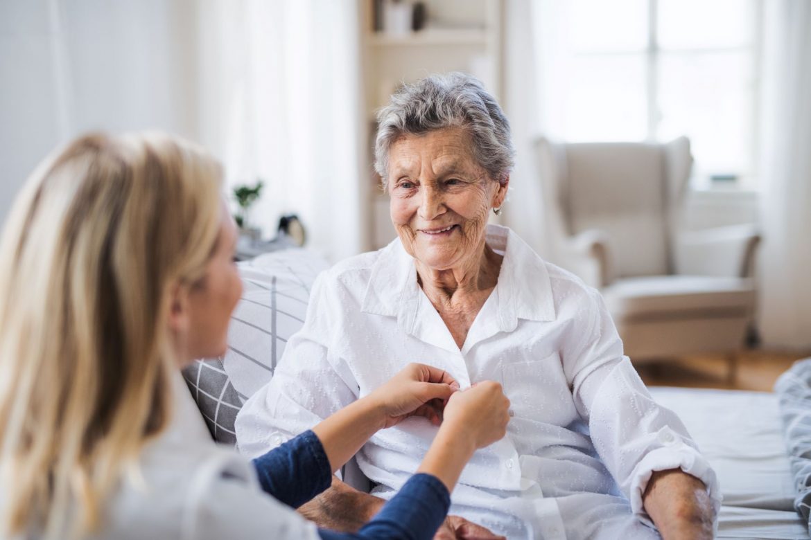 Objawy Alzheimera - jak rozpoznać chorobę Alzheimera? Lekarka zapina seniorce koszulę na guziki na wizycie domowej.