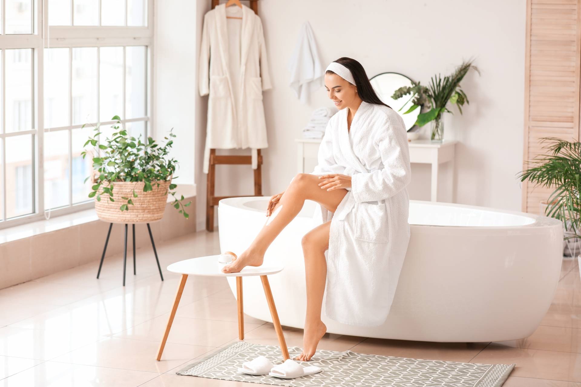 Czym jest cellulit i jak z nim walczyć? Co jeść, jak ćwiczyć i jakie zabiegi zastosować? Kobieta w szlafroku siedzi na krawędzi wanny w łazience i wsmarowuje balsam w łydkę.