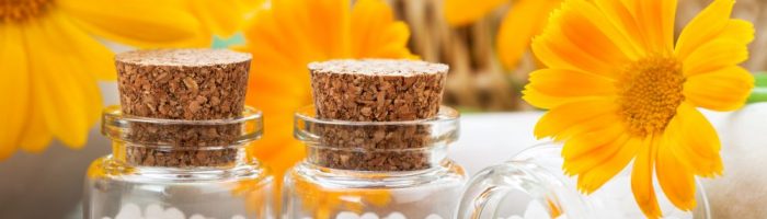 Jaka jest różnica między lekiem homeopatycznym pojedynczym a złożonym? Leki homeopatyczne w granulkach w słoiczkach w otoczeniu pomarańczowych kwiatów.