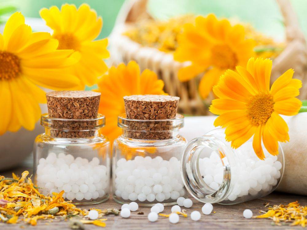 Jaka jest różnica między lekiem homeopatycznym pojedynczym a złożonym? Leki homeopatyczne w granulkach w słoiczkach w otoczeniu pomarańczowych kwiatów.