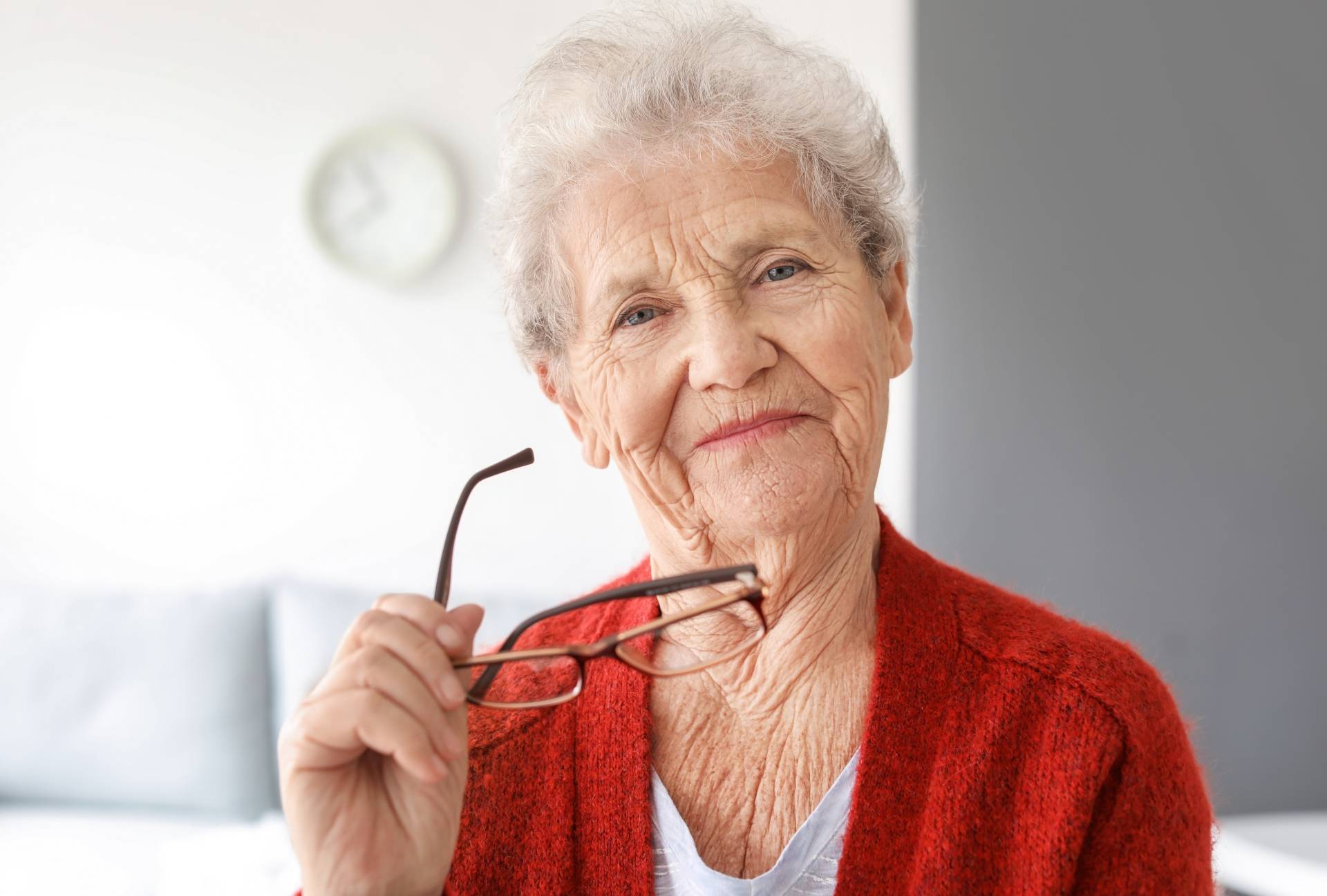 Jaskra - jak rozpoznać i leczyć chorobę oczu? Uśmiechnięta seniorka w domu trzyma okulary w ręku i patrzy w obiektyw.