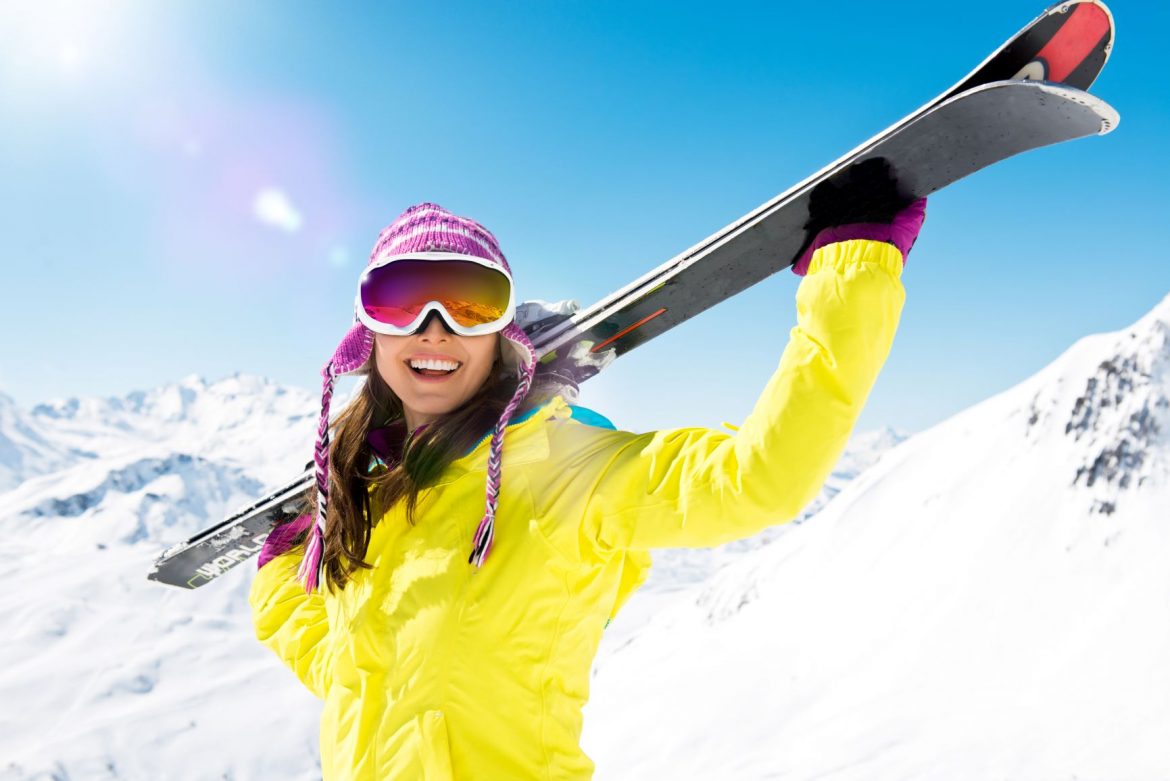 Jazda na nartach - jak się przygotować? Uśmiechnięta kobieta w żółtym kombinezonie trzyma w rękach narty na stoku górskim.