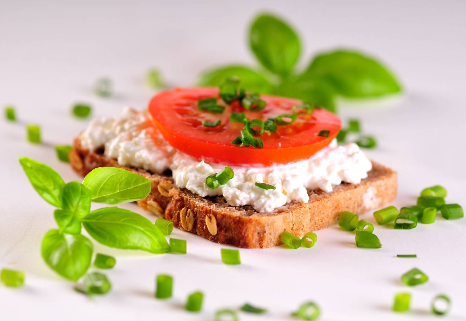 Dieta na odporność - unikaj produktów zawierających gluten. Kromka ciemnego pieczywa z twarożkiem, pomidorem, szczypiorkiem i listkami bazylii leży na białym blacie.
