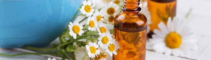 Naturalne antybiotyki, czyli leczenie roślinami i ziołami. Kwiatki rumianku leżą na rustykalnej drewnianej podłodze, obok stoi niebieska miska z rumiankową kąpielą i dwie brązowe buteleczki z syropem.