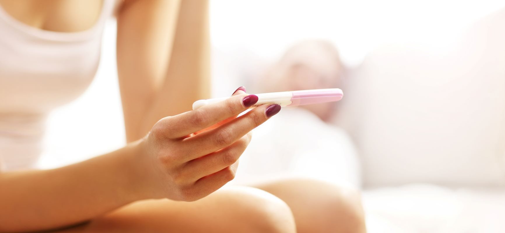 Kiedy zrobić test ciążowy, aby jego wynik był wiarygodny? Kobieta w białym topie i majtkach siedzi na łóżku i czeka na wynik testu ciążowego.