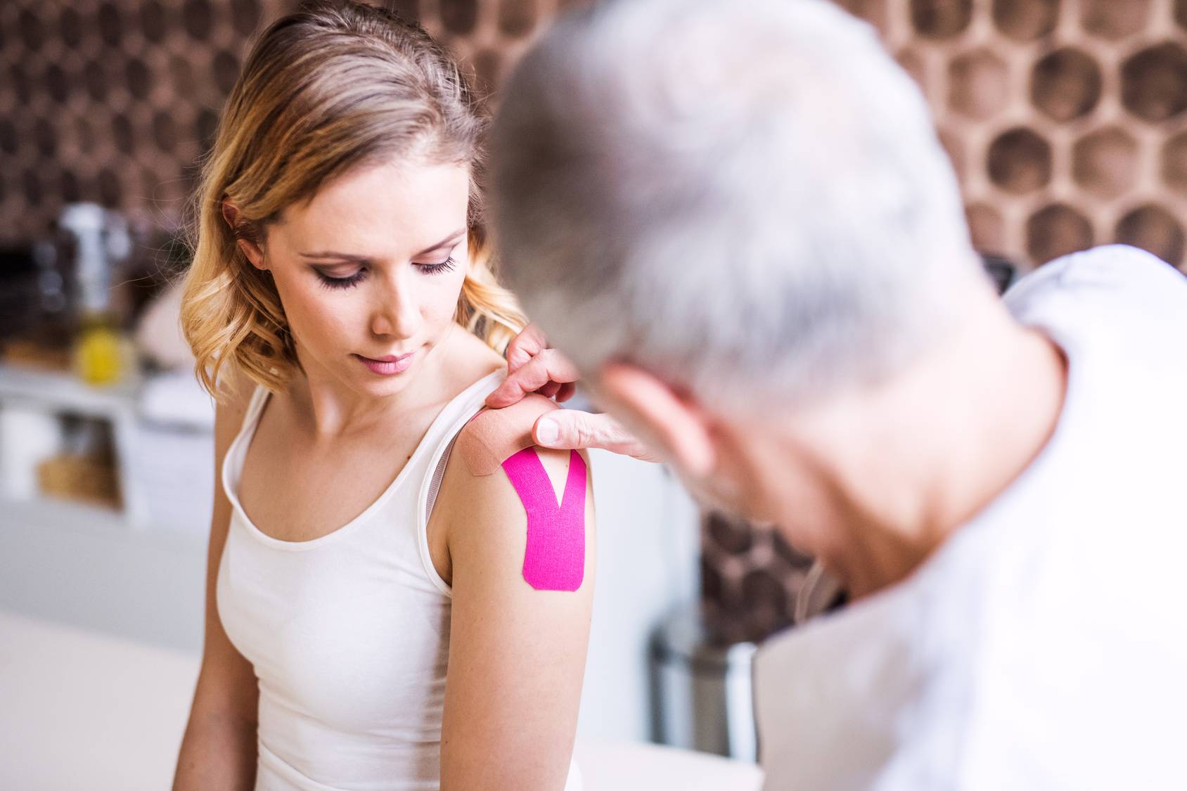 Kinesiotaping - taśma kinezjologiczna na urazy. Fizjoterapeuta przykleja pacjentce tejpy na ramię.