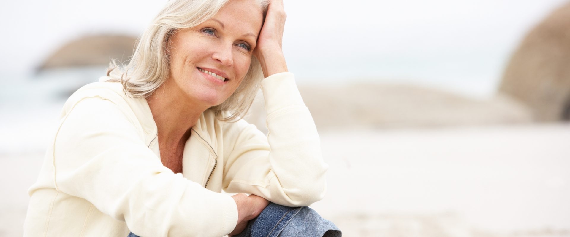 Kintsugi w psychoterapii: szanuj swoje blizny. Kobieta w średnim wieku w jasnym swetrze i podwiniętych jeansach siedzi na plaży. Jest lekko uśmiechnięta i zamyślona.