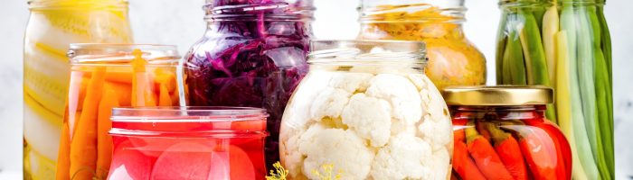 Mikroflora jelitowa - jak dbać o jej równowagę? Kiszonki z kolorowych warzyw w słoikach stoją na rustykalnym drewnianym stole.