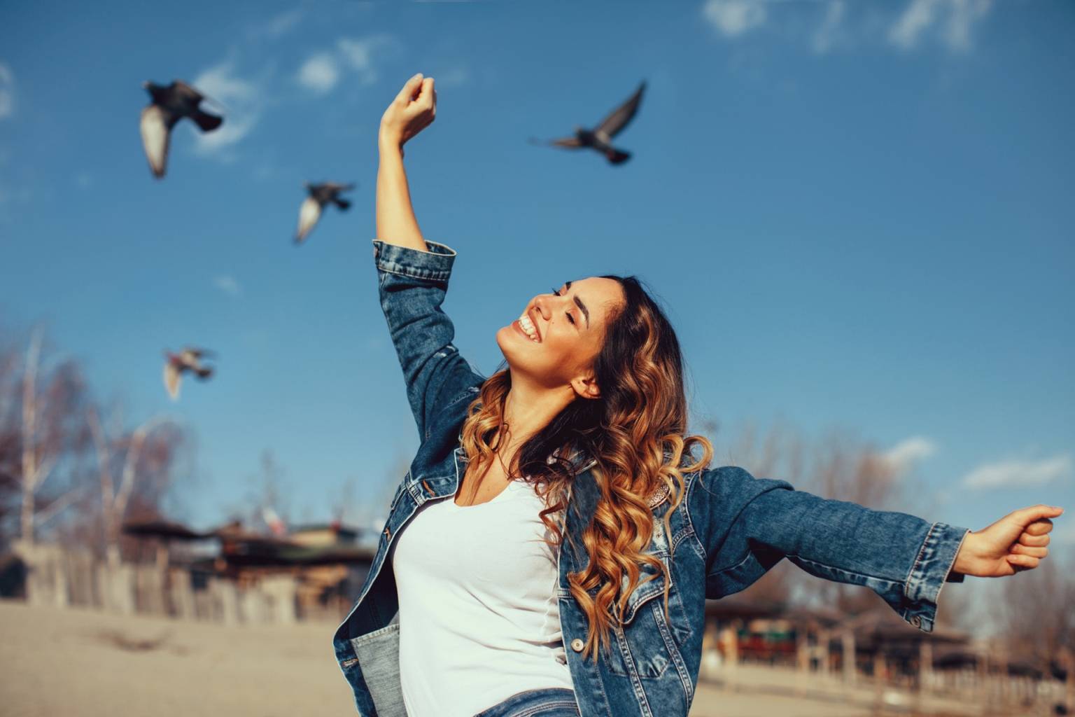Czy sztuczne światło nam szkodzi? Szczęśliwa uśmiechnięta dziewczyna w białym topie i jeansowej kurtce cieszy się piękną pogodą, w tle latają ptaki.