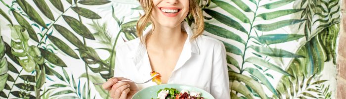 Co zrobić, żeby schudnąć? Uśmiechnięta kobieta, trzymająca talerz ze zdrowym jedzeniem, stoi na tle ściany powleczonej piękną tapetą w zielone egzotyczne liście.