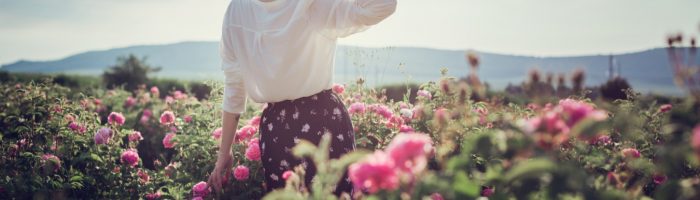 Zaburzenia miesiączkowania - co jest ich przyczyną według medycyny chińskiej? Młoda dziewczyna w białej bluzce i słomkowym kapeluszu stoi na łące wśród różowych kwiatów.