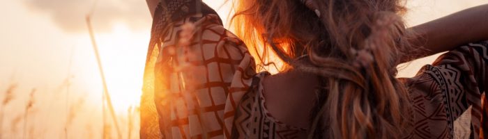 Lek homeopatyczny Staphysagria pomaga przy dolegliwościach psychosomatycznych i uwalnia emocje. Kobieta w zwiewnej letniej sukience stoi na łące tyłem do aparatu i podziwia zachodzące słońce.