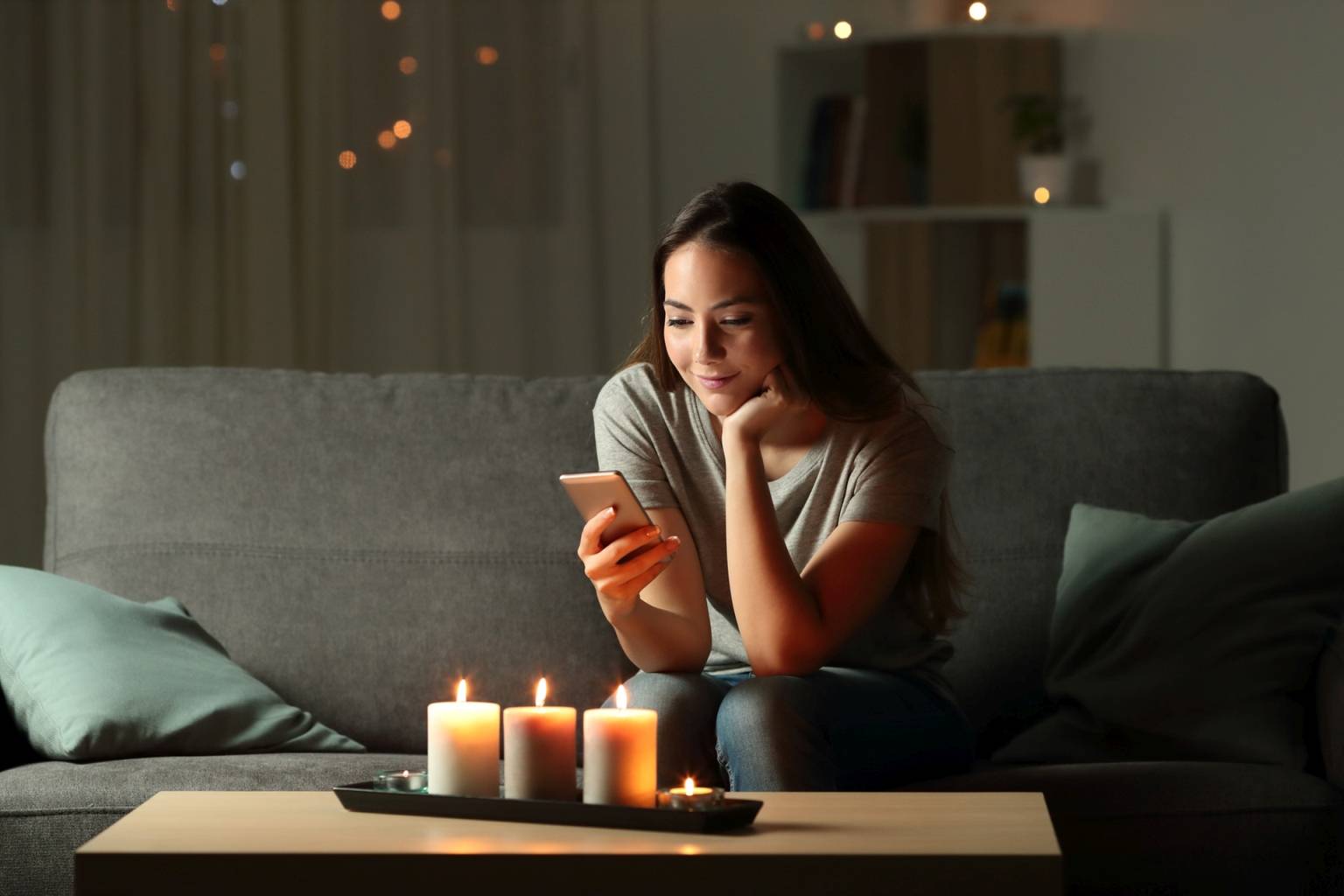 Czy sztuczne światło wpływa na nasze życie i zdrowie? Kobieta siedzi wieczorem na kanapie w salonie przy świecach i przegląda smartfona.