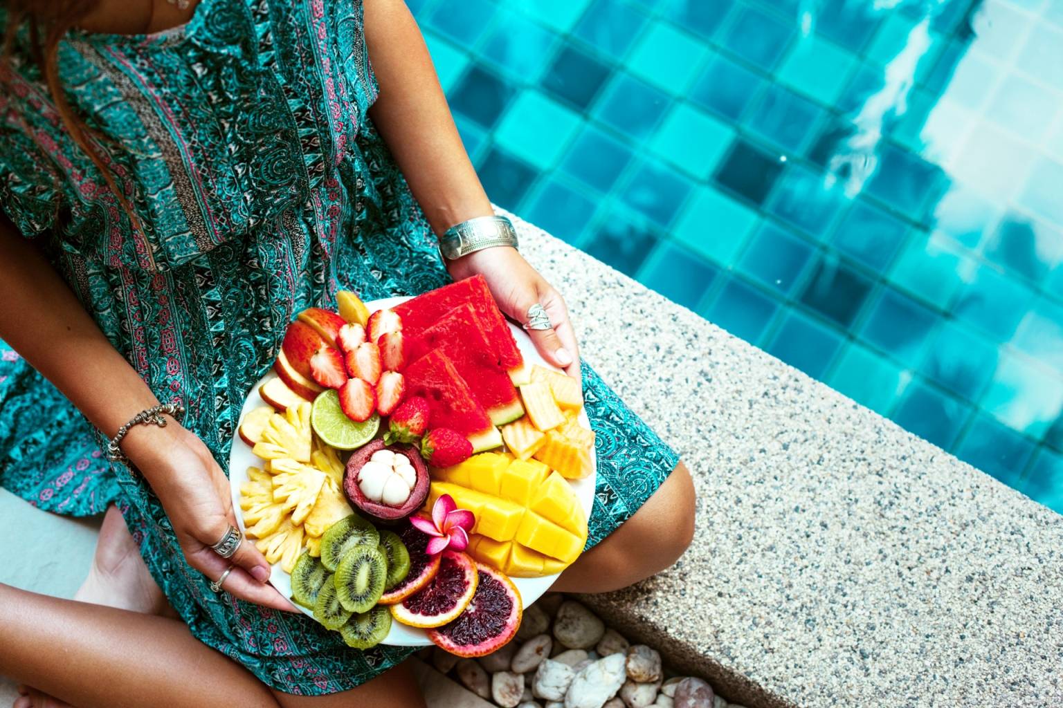 Nietolerancja fruktozy - dlaczego owoce szkodzą? Opalona dziewczyna w niebieskiej letniej sukience siedzi nad basenem i trzyma w rękach talerz z egzotycznymi owocami.