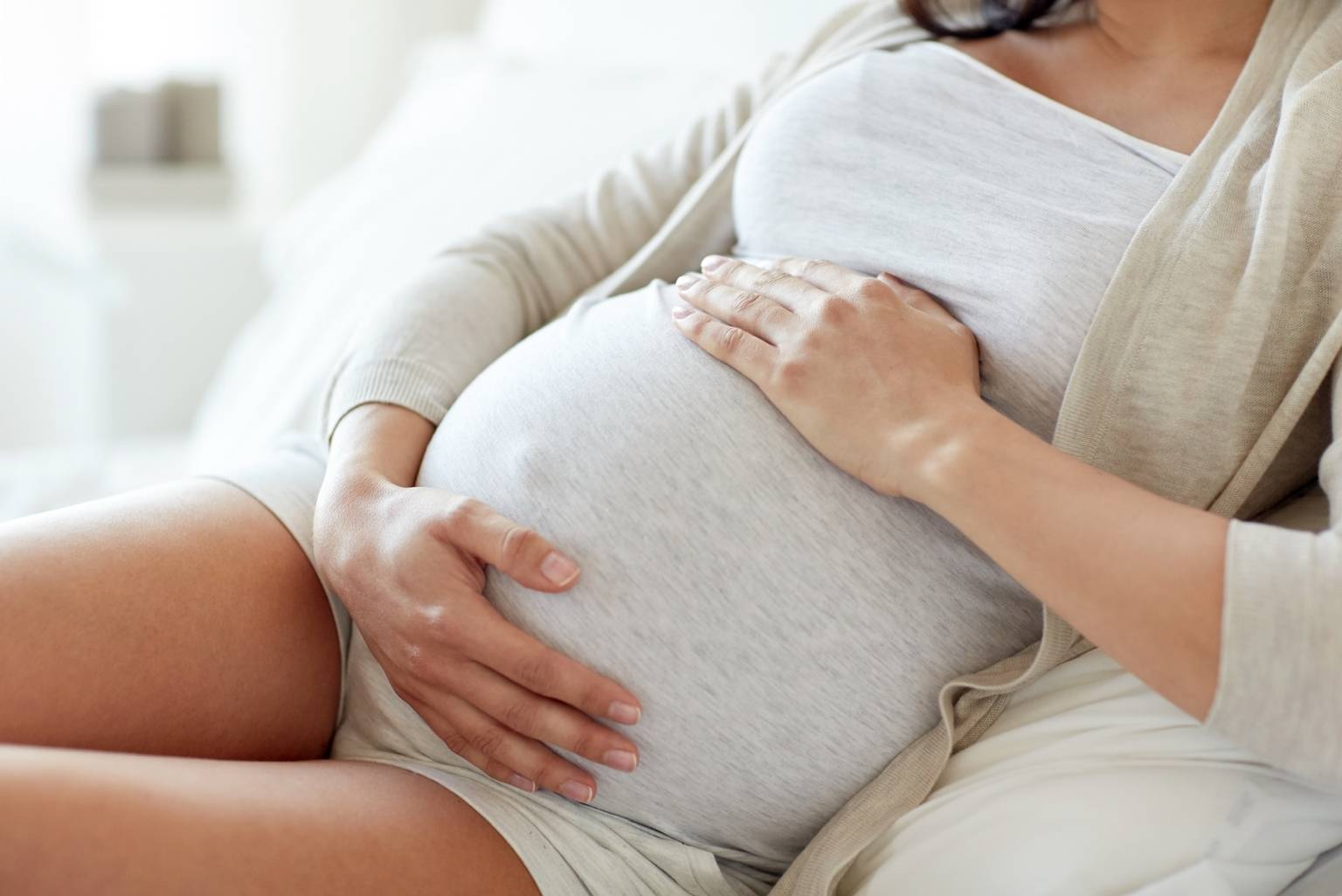 Leczenie niepłodności w medycynie chińskiej. Kobieta w zaawansowanej ciąży w szarej bluzce, narzutce i krótkich spodenkach siedzi na kanapie i trzyma się za brzuch. 