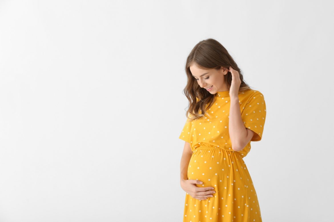 Nudności w ciąży - jak radzić sobie z mdłościami w ciąży? Piękna kobieta w ciąży w żółtej sukience na tle białej ściany.