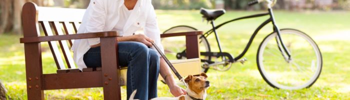 Homeopatia na menopauzę i okres przekwitania. Jakie są objawy klimakterium? Dojrzała kobieta siedzi na ławce w parku i głaszcze swojego psa, w tle stoi jej rower.
