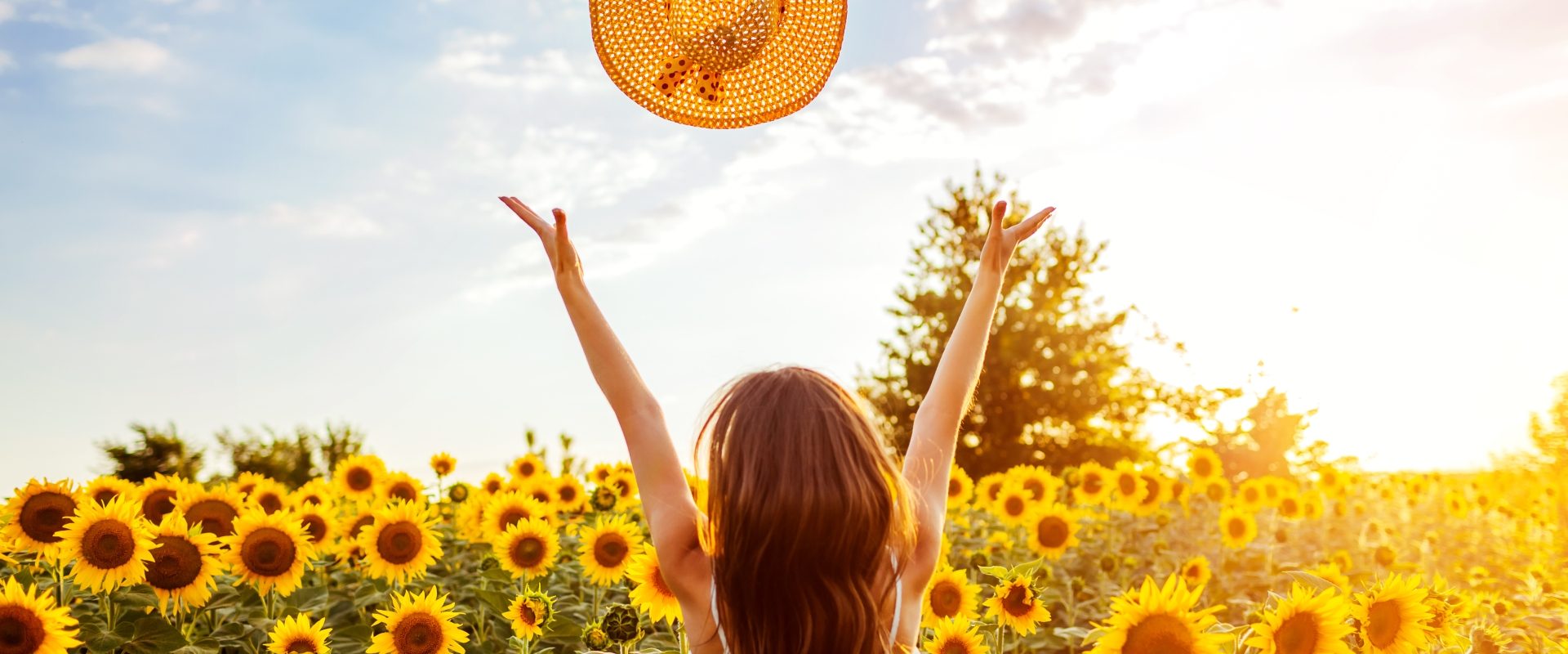 Ikigai - jak pokochać codzienność? Długowłosa kobieta w polu słoneczników podrzuca słomkowy kapelusz w geście radości.