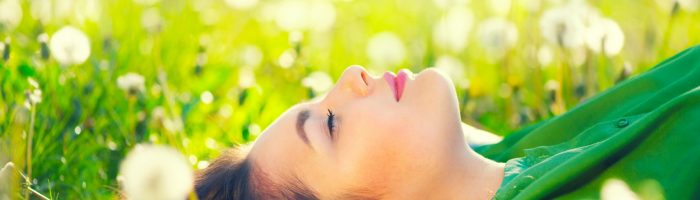 Kalendarz pylenia roślin - alergia na pyłki. Młoda, zamyślona i szczęśliwa kobieta w zielonej bluzce leży na trawie wśróddmuchawców.