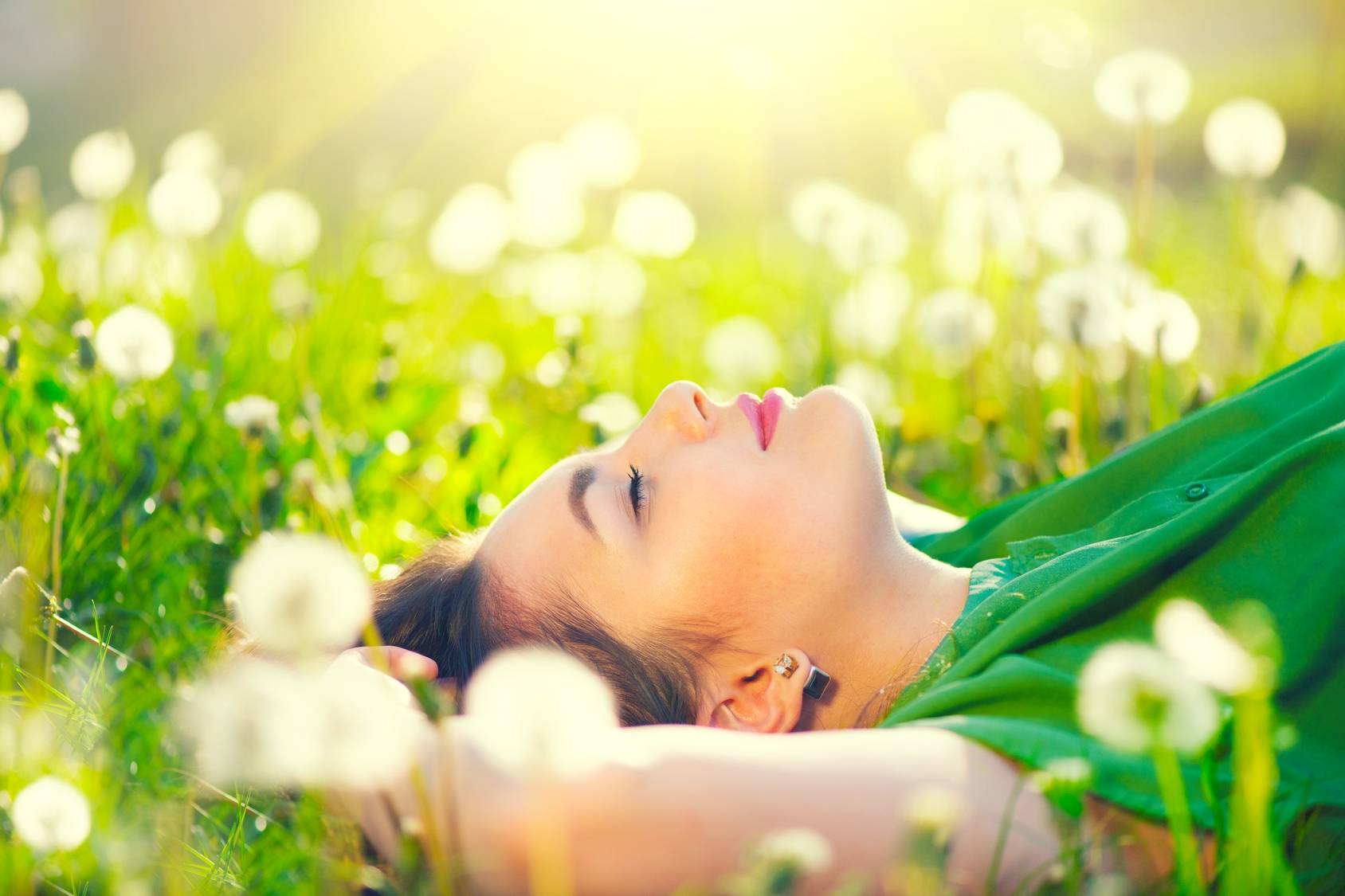 Kalendarz pylenia roślin - alergia na pyłki. Młoda, zamyślona i szczęśliwa kobieta w zielonej bluzce leży na trawie wśróddmuchawców.