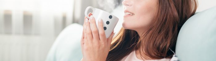 Homeopatia i leki homeopatyczne na ból gardła - jak leczyć zapalenie gardła? Kobieta siedzi na kanapie i pije gorącą herbatę.