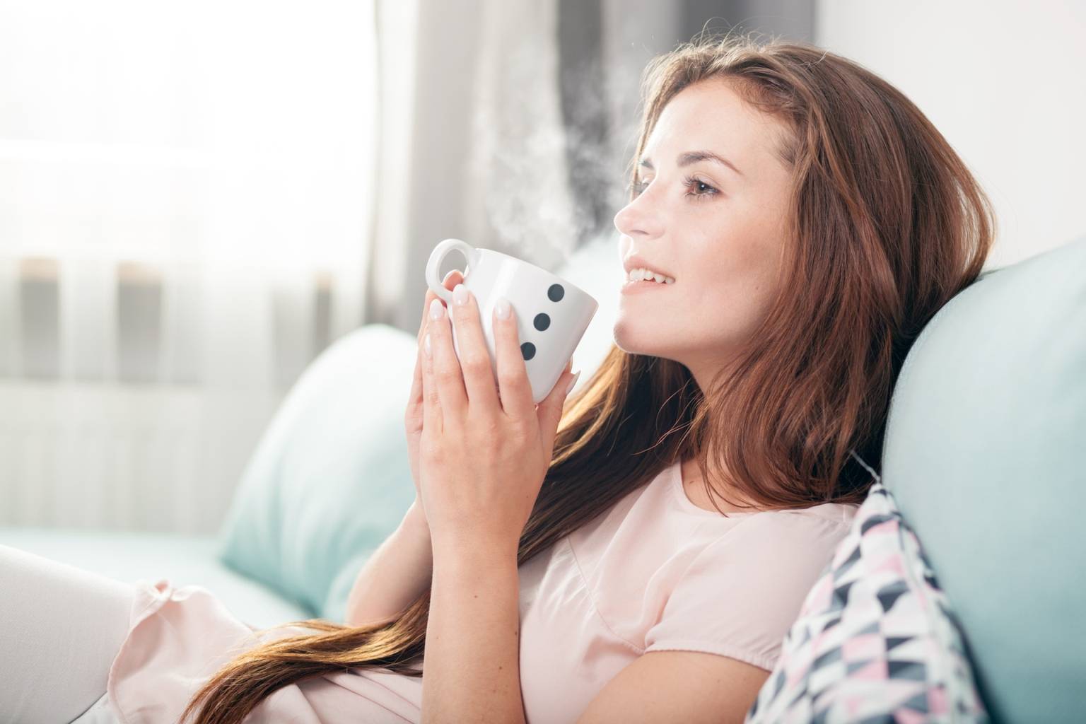 Homeopatia i leki homeopatyczne na ból gardła - jak leczyć zapalenie gardła? Kobieta siedzi na kanapie i pije gorącą herbatę.
