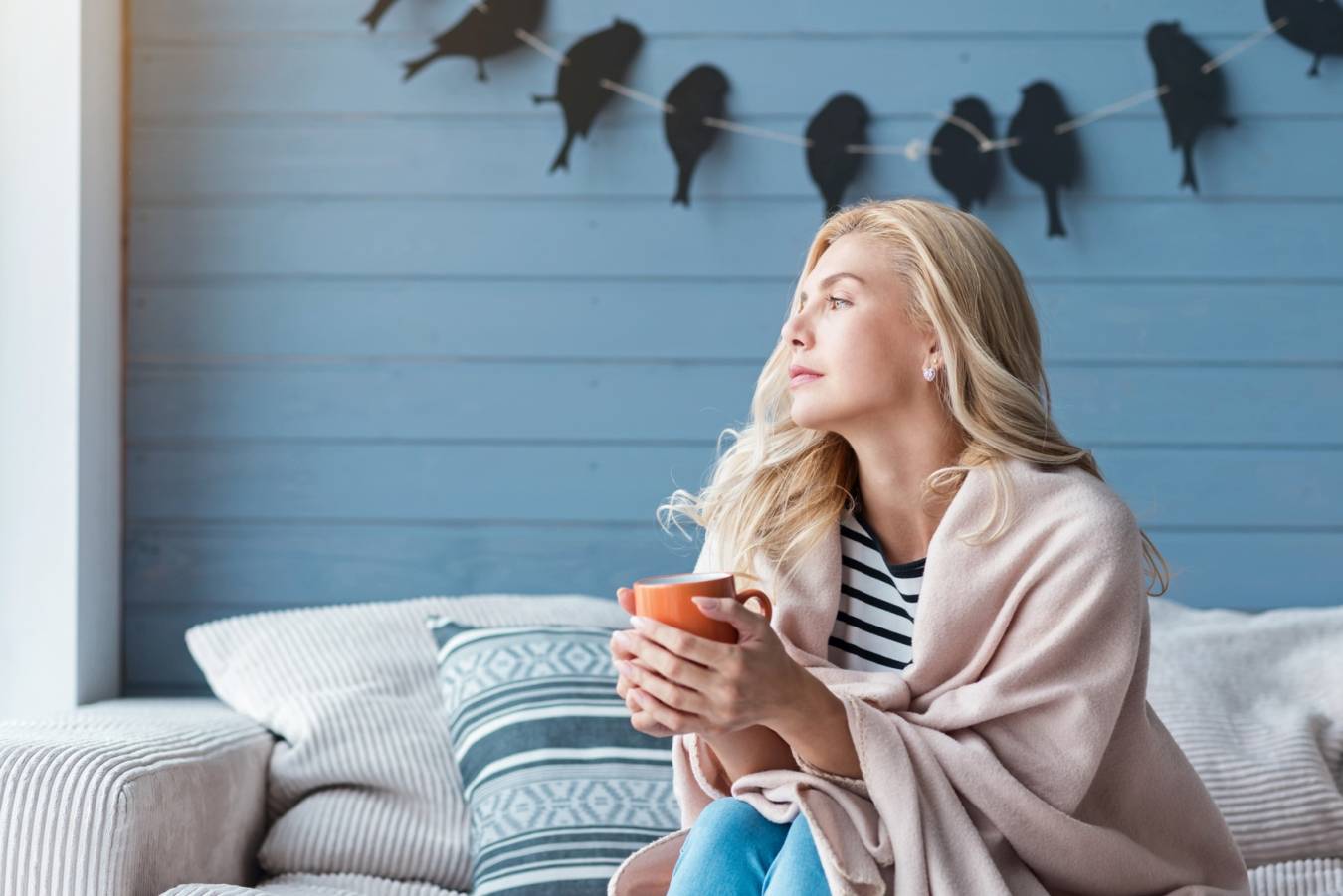 Jakie są objawy grypy? Jak leczyć grypę? Blondwłosa kobieta siedzi na kanapie opatulona kocem i pije gorącą herbatę.