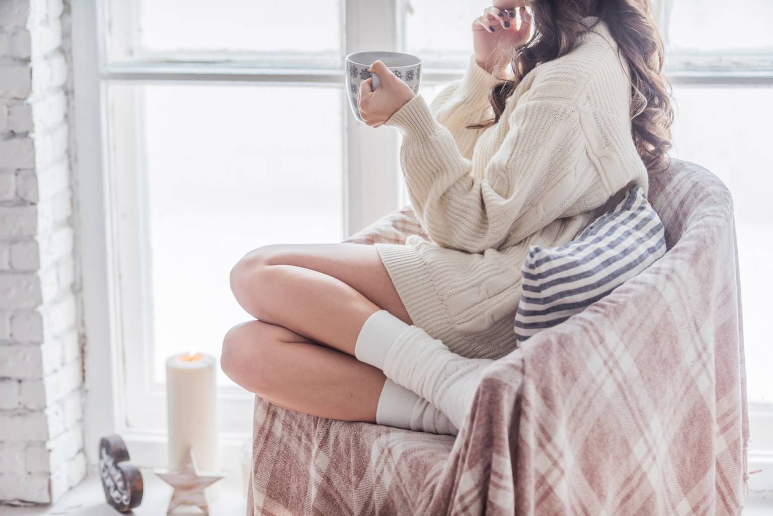 Jak szybko wyleczyć przeziębienie? Co na przeziębienie? Kobieta siedzi na fotelu w kocu, z podkulonymi nogami, w ciepłym swetrze i skarpetkach, pije gorącą herbatę i patrzy w okno.