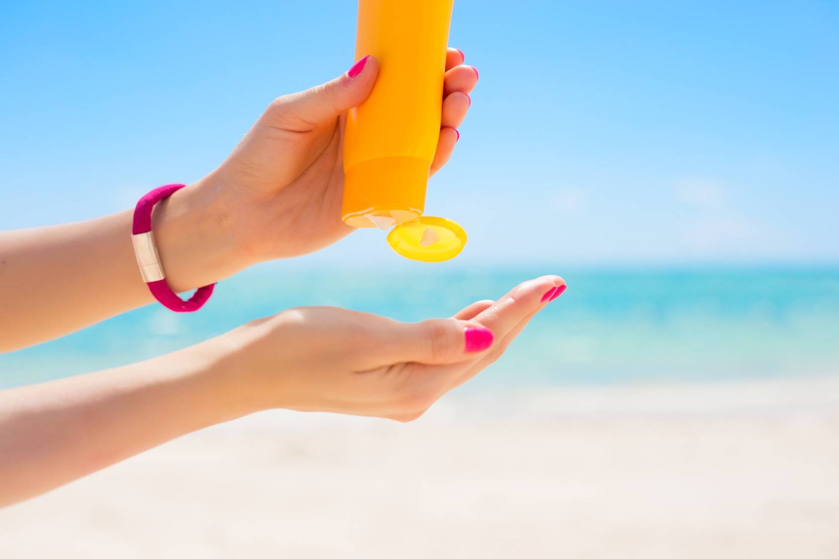 Jak czytać oznaczenia na kremach przeciwsłonecznych i jak wybrać bezpieczne filtry UV? Zbliżenie na kobiece ręce nakładające porcję kremu z żółtego opakowania na plaży.