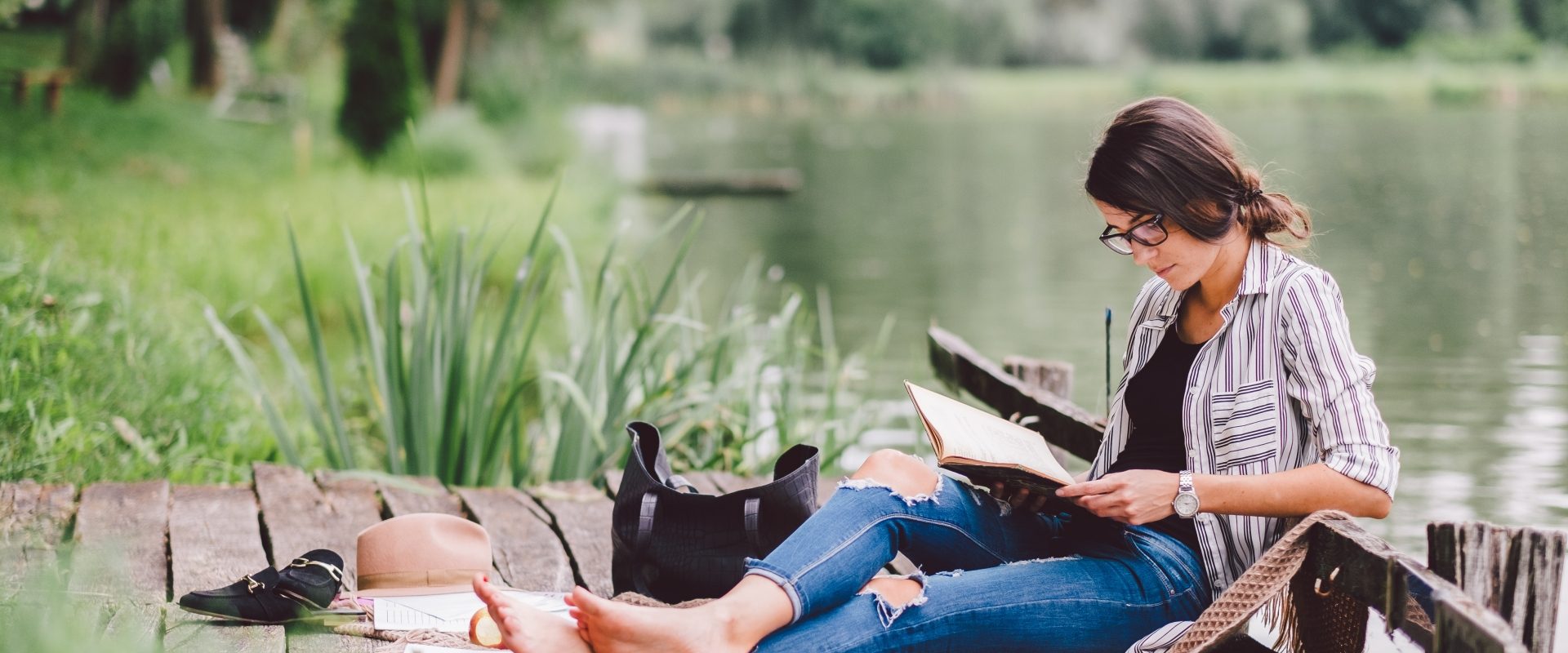 Książki na lato - kobieta w okularach w rogowej oprawce czyta książkę na pomoście nad jeziorem.