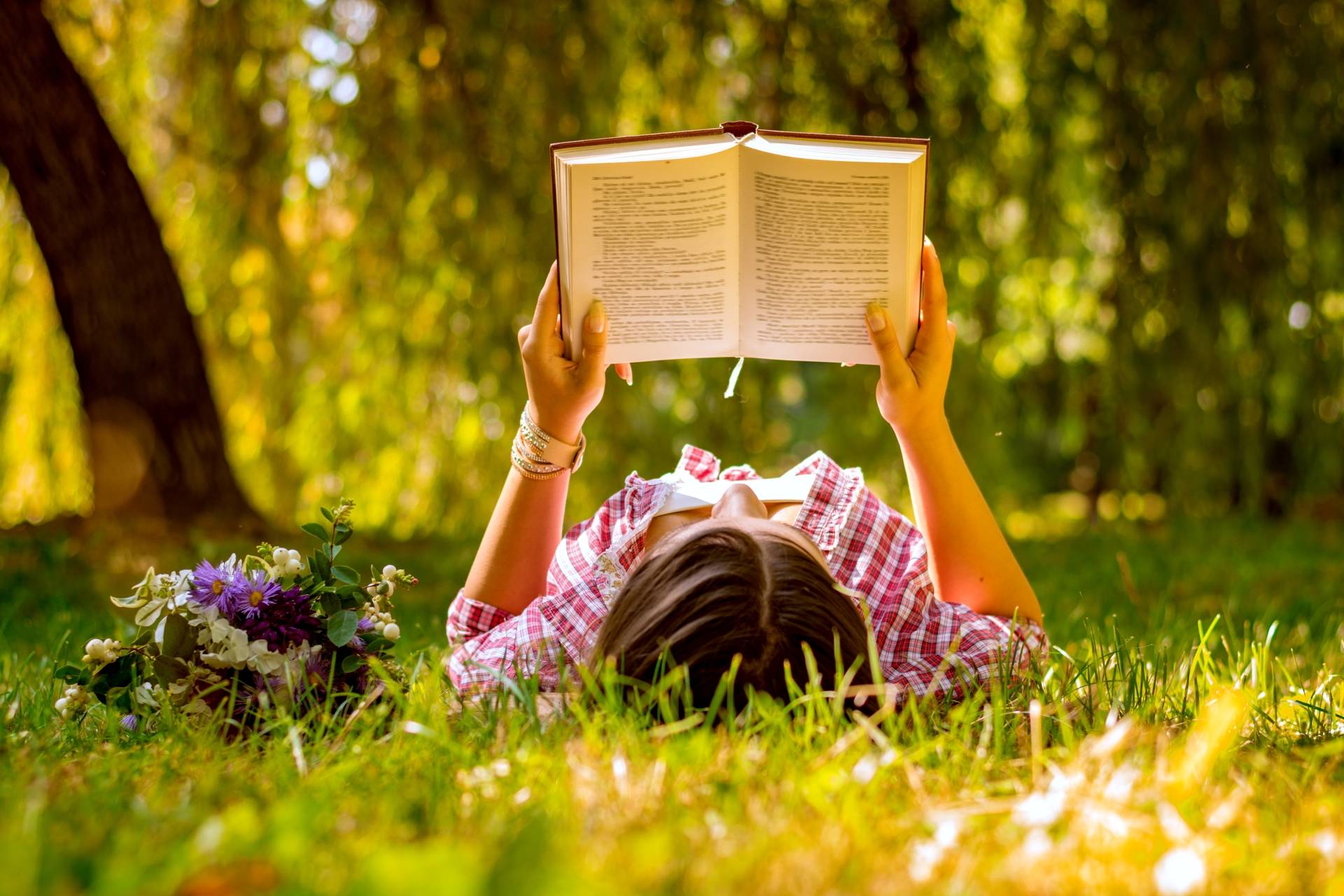 Książki na wakacje - kobieta czyta książkę, leżąc na trawie w popołudniowym słońcu.